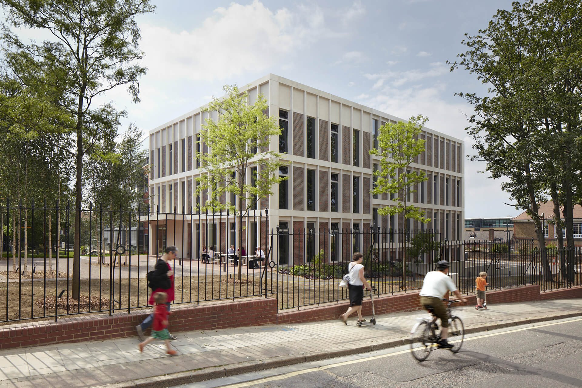 Three-storey building with floor-to-ceiling windows, with people sitting at tables in front of it. Around the house is a courtyard with trees, the house is surrounded by a fence, in front of it people are walking on the pavement and the street. 