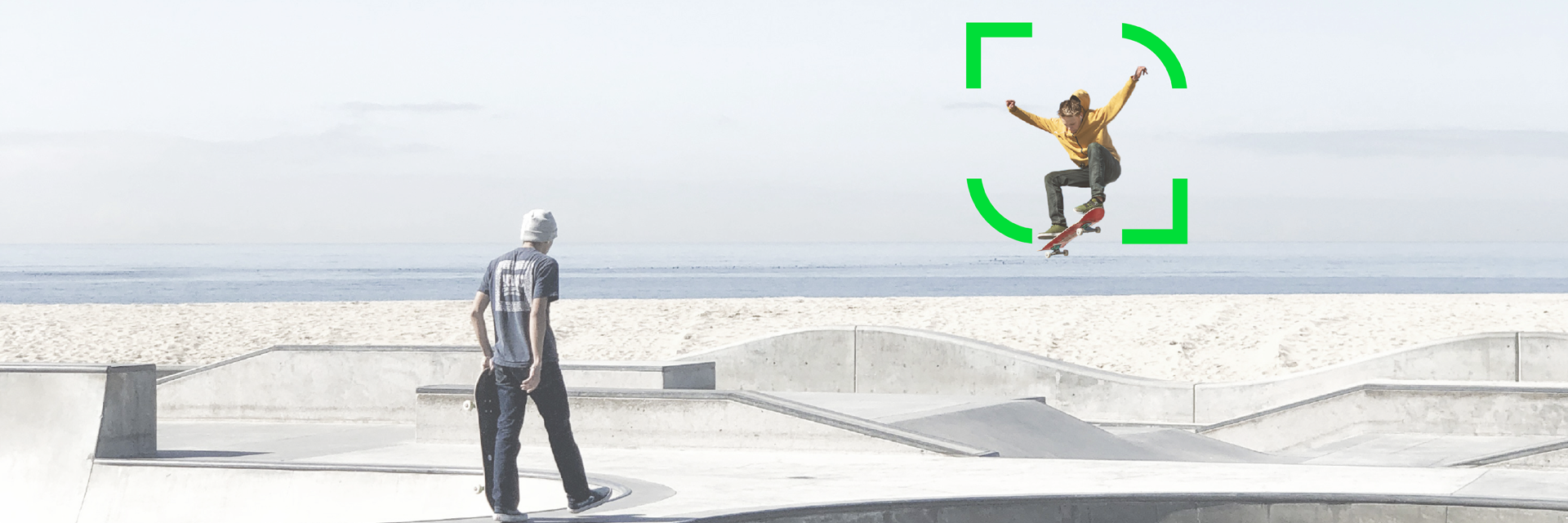 Two people with skateboards in a concrete skate park, one is doing a jump, sea in the background