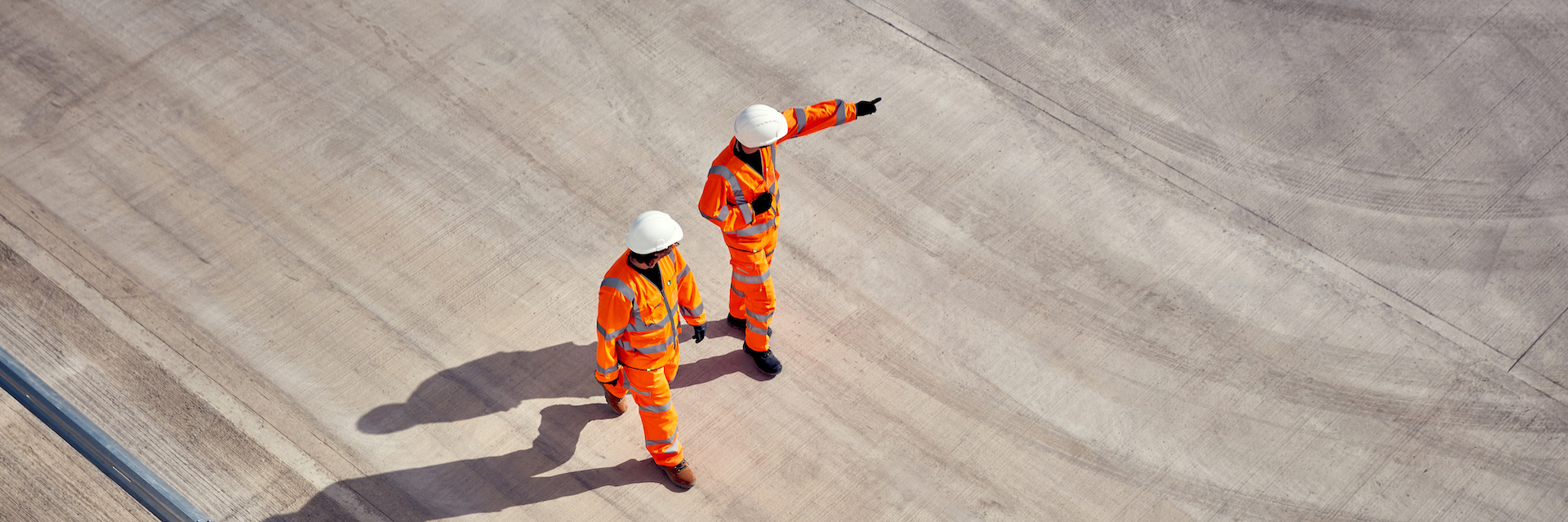 Two construction workers in orange high-visibility clothing and helmets stand on a large concrete surface; one points forward while the other observes, suggesting direction on a works site.