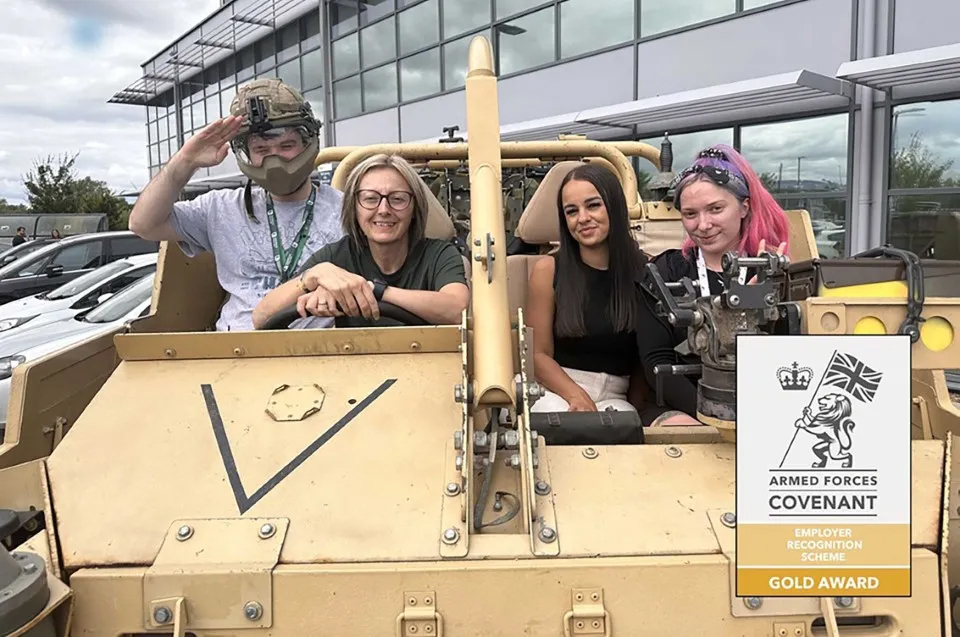 Four people seated in a tan military vehicle, with one person saluting while wearing camo gear. The vehicle is displayed outdoors near a modern glass building. A sign on the vehicle's front corner shows the Armed Forces Covenant Gold Award emblem.