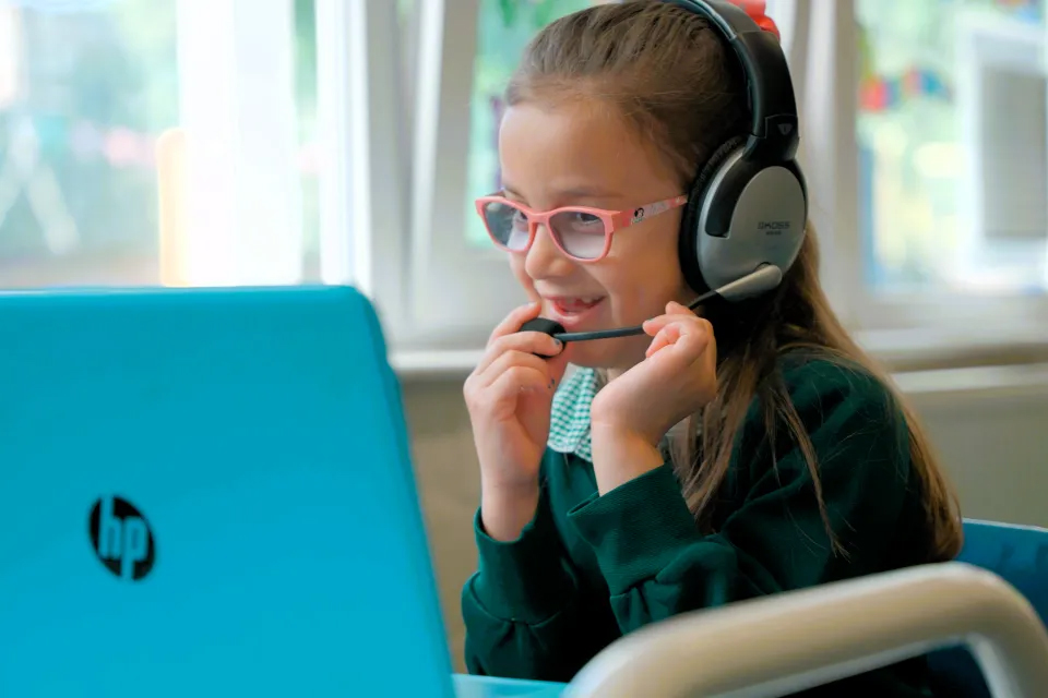 A child wearing headphones sits at a desk using a blue HP laptop in a bright classroom with large windows in the background.
