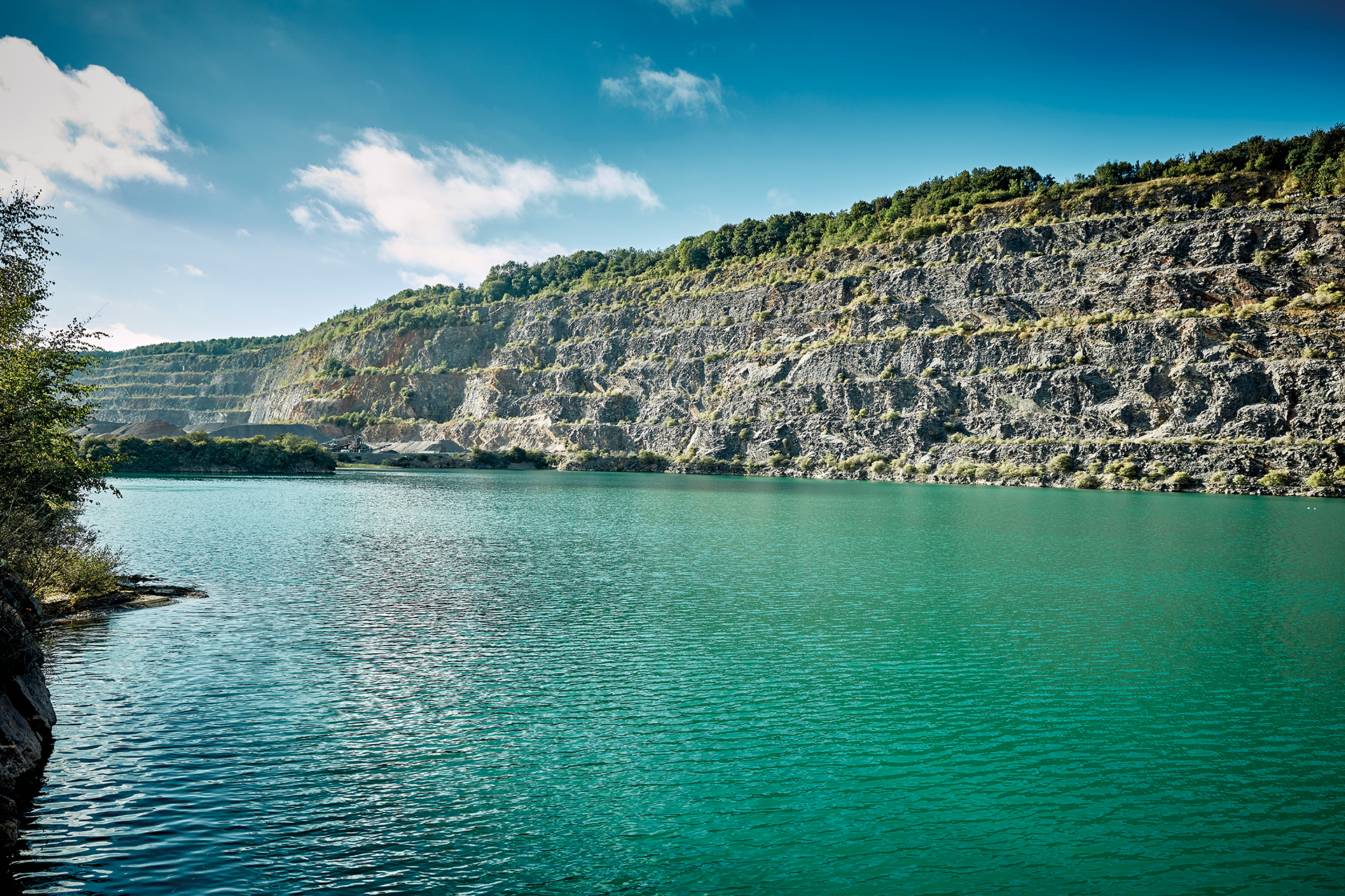 Turquoise lake surrounded by steep rocky cliffs, with greenery along the ridge and trees on the left edge under a partly cloudy blue sky.