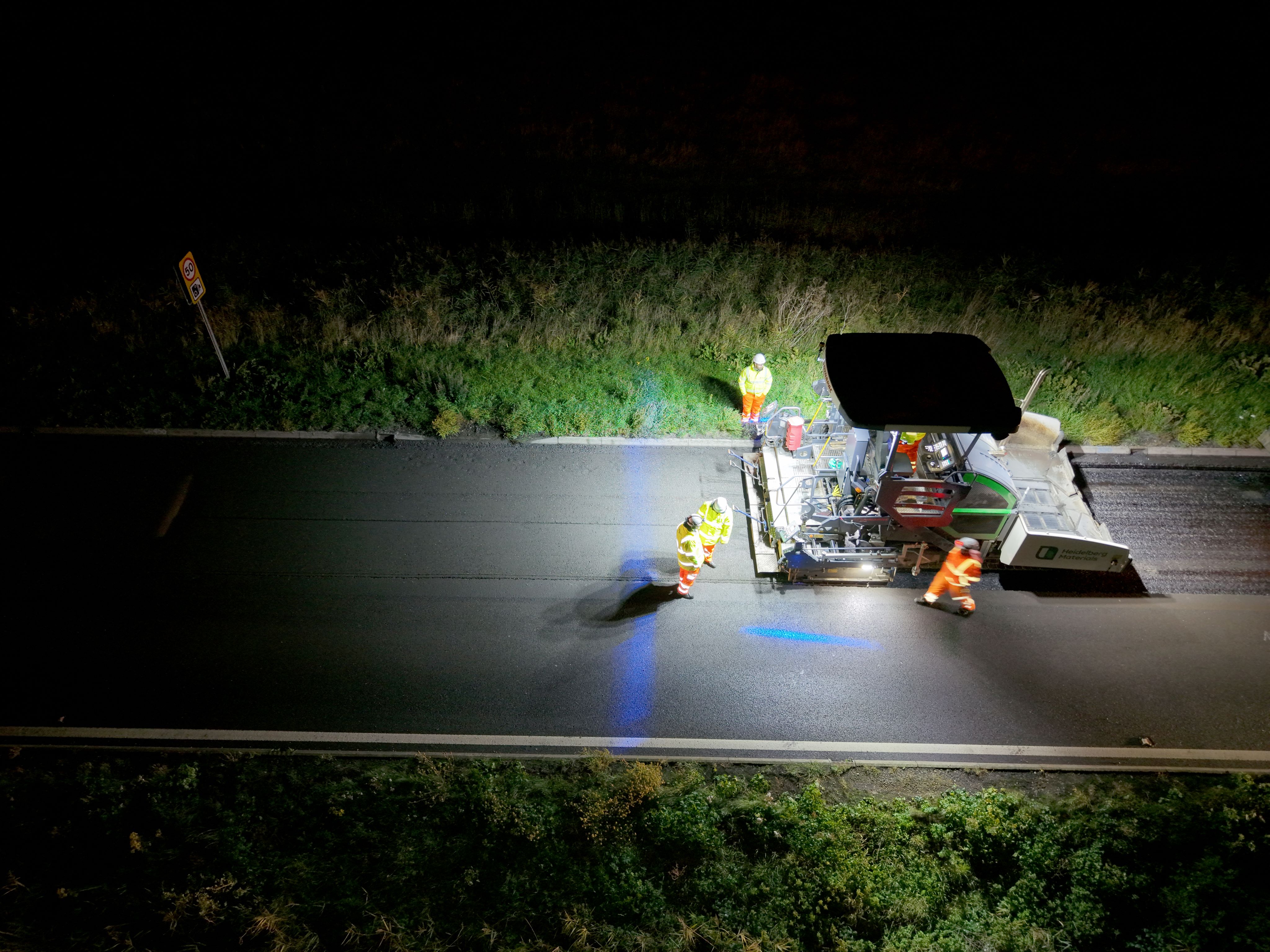 Construction workers operate machinery on a road at night under bright lighting.