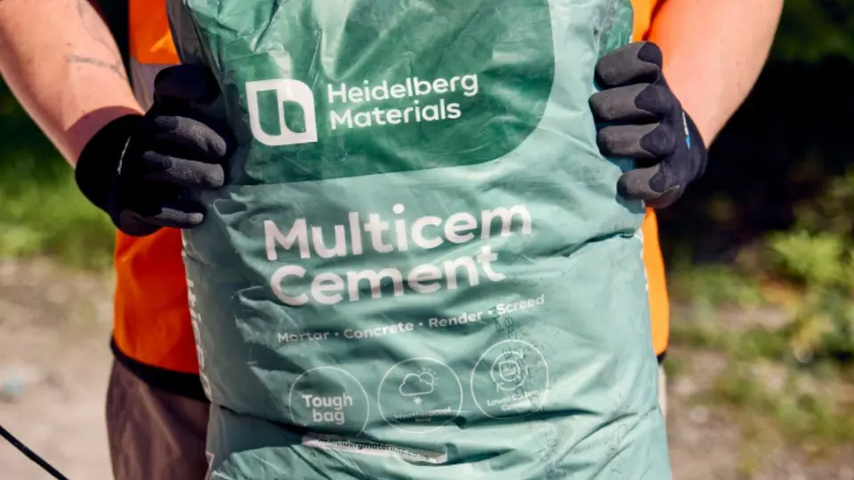 A worker holding a green Heidelberg Materials Multicem Cement bag outdoors.