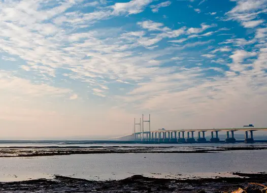 Severn Bridge spanning across a wide river estuary under a partly cloudy blue sky, with muddy shoreline and sparse vegetation in the foreground.