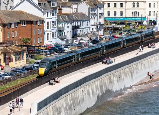 Coastal town with a train traveling along a track parallel to the shoreline. A sandy beach with scattered visitors lies to the right, bordered by the sea. On the left, colorful houses and buildings line the town, with parked cars visible along the road.